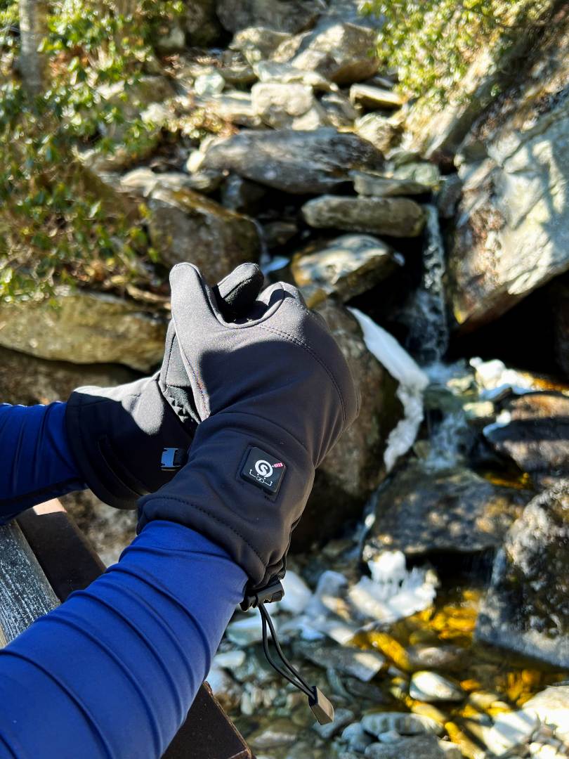 A person wearing black heated gloves near a creek in a natural outdoor setting. The gloves have a visible control button on the wrist, and the person is adjusting them as they stand beside rocks and flowing water in a scenic mountainous environment