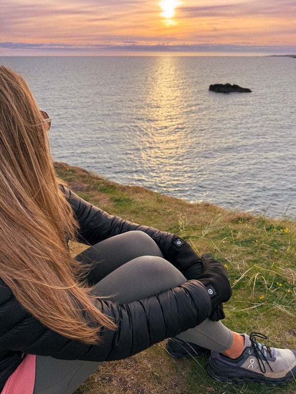 A person wearing heated gloves sits on a grassy cliff, watching the sunset over the ocean, dressed in a black jacket, gray leggings, and sneakers