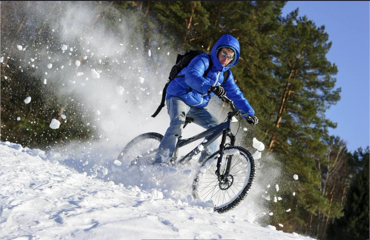 A cyclist in a blue jacket and helmet riding through snow, with snow flying as the bike tires cut through the terrain