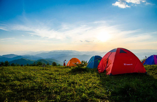 Camping tents set up on a grassy hilltop with a scenic mountain view at sunset. The colorful tents are surrounded by nature, offering a perfect outdoor experience