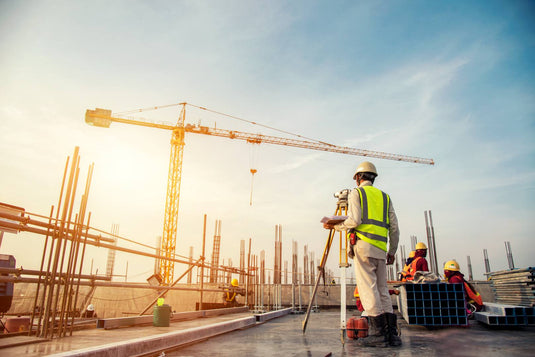 Construction worker surveying the site with a level tool, with a crane and workers in the background at a construction site.