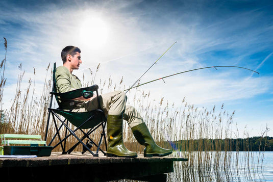 A person fishing by a lake, sitting on a chair with a fishing rod, wearing green boots and surrounded by tall grass.