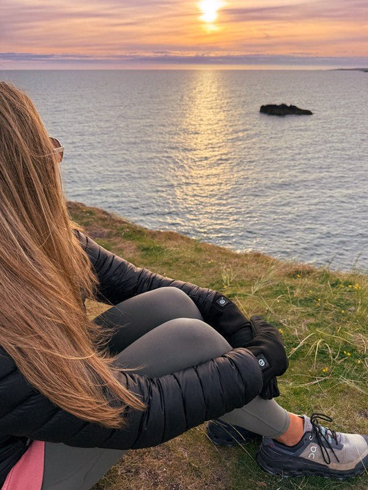 A person wearing heated gloves sits on a grassy cliff, watching the sunset over the ocean, dressed in a black jacket, gray leggings, and sneakers