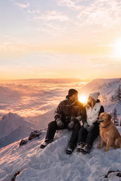 Couple with a dog on a snowy mountain peak at sunset in a winter landscape